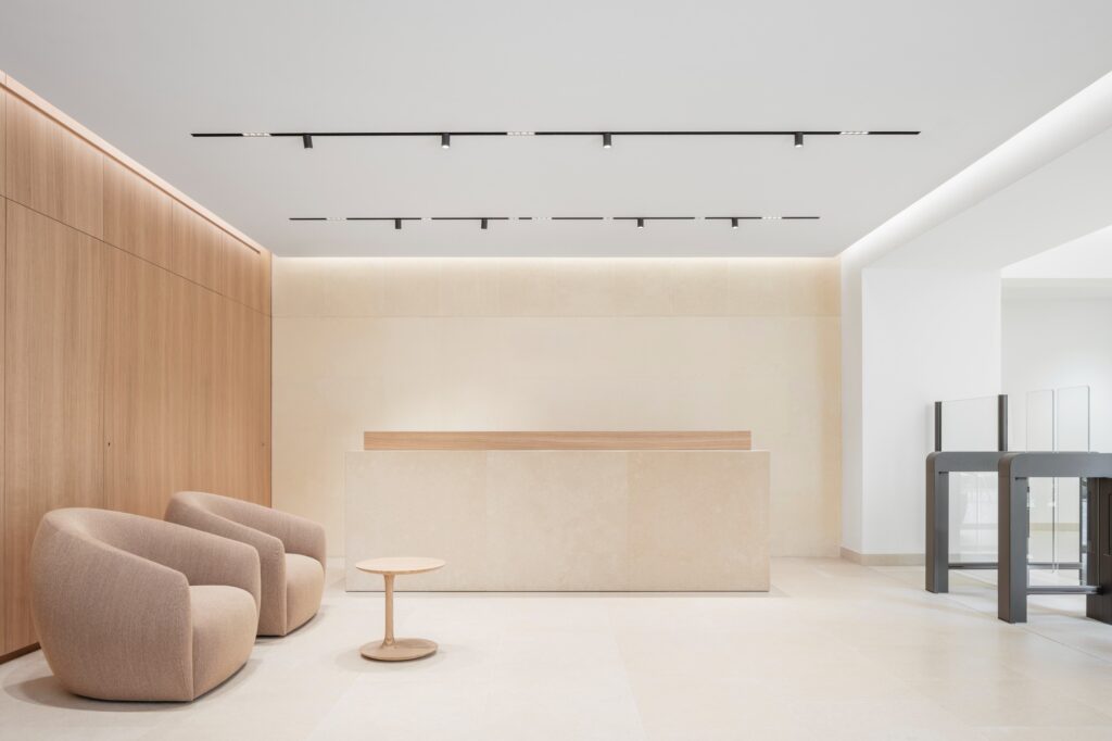 A minimalist reception area with a stone desk, wooden wall paneling, and two neutral-toned lounge chairs.