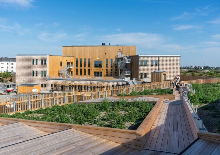 A wide view of Ängsdal School featuring a mix of wooden facades, patterned concrete walls, and extensive outdoor learning decks with green landscaping.