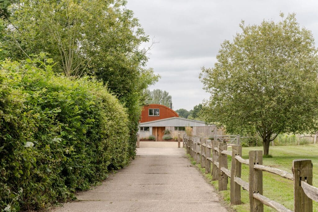A long driveway leading to the Dutch Barn house framed by tall hedges and a rustic wooden fence.