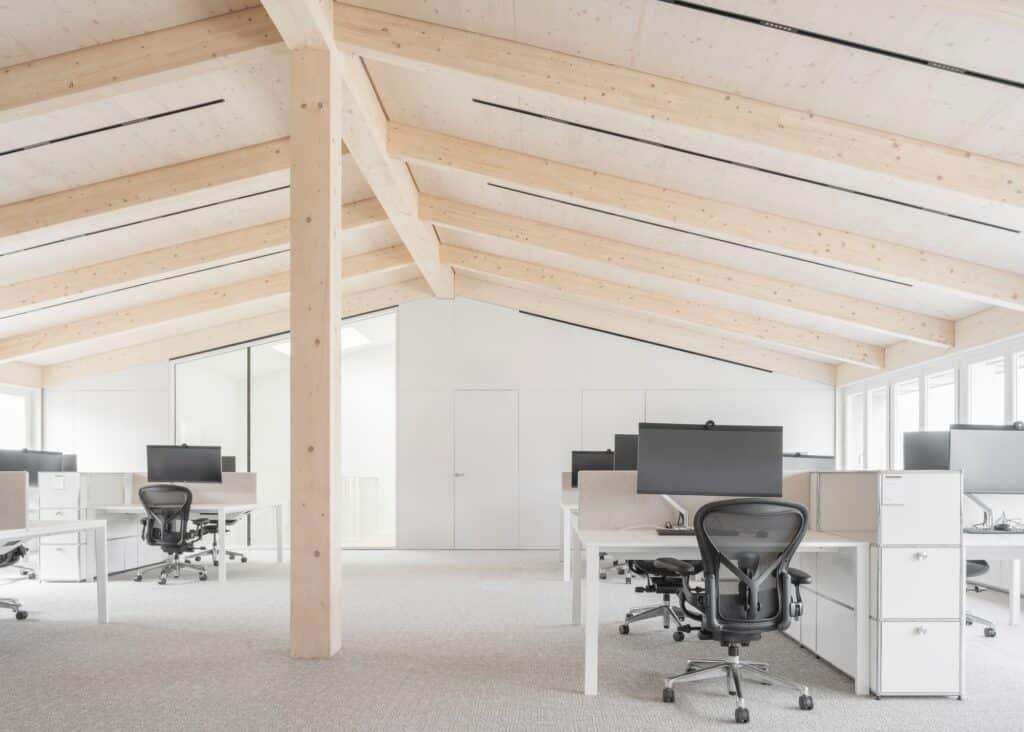 A large open-plan office space under a vaulted timber roof structure with modern workstations and black ergonomic chairs.