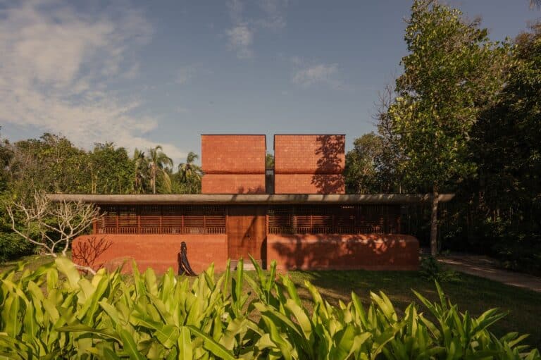 Sumesh House in Payyanur: Architectural composition featuring exposed laterite brick walls, a long cantilevered roof, and timber screens, seamlessly integrating the cultural context with the surrounding lush tropical nature.
