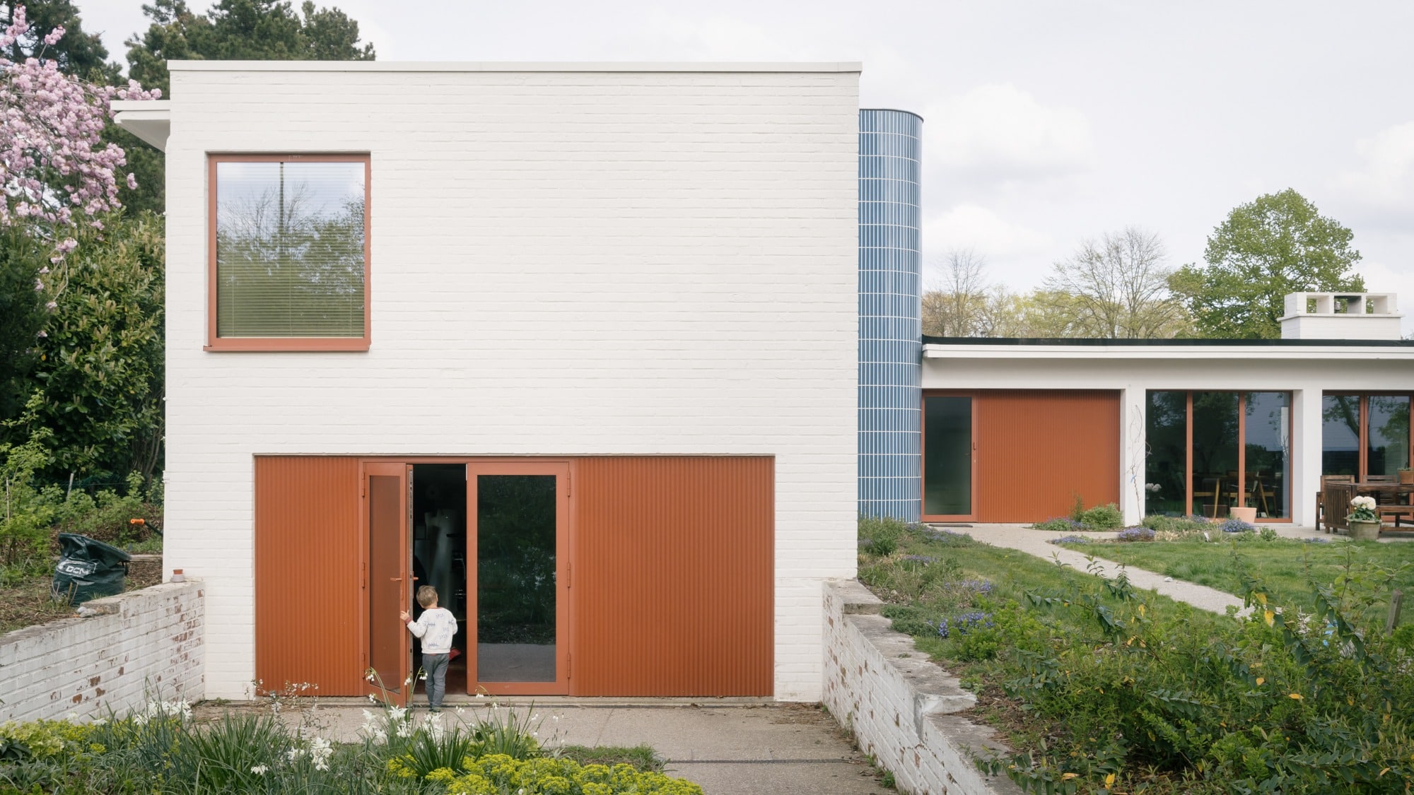 Modern bungalow exterior featuring white brick walls, a blue tiled cylindrical tower, and orange sliding doors with a child standing at the entrance.