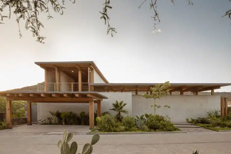 Front facade of Casa Altozano in Queretaro with concrete walls, wooden roof structures, and lush desert landscaping.
