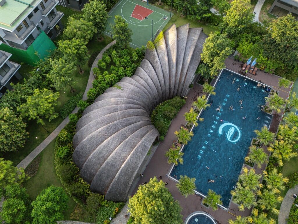 Aerial view of Goot Garden House showing the organic shell-like roof structure adjacent to a blue swimming pool and surrounding greenery in Eco Central Park.