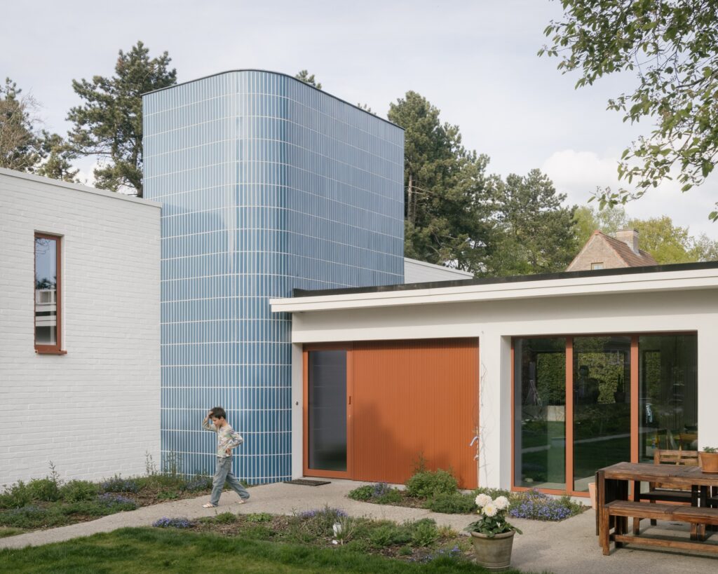 Modern bungalow exterior with white brick walls, a blue tiled tower, orange sliding doors, and a child standing at the entrance.