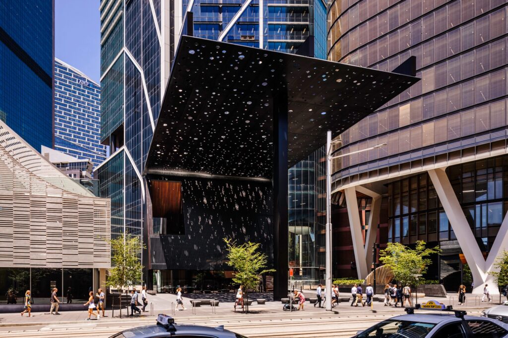 Street view of the George Street Plaza showing the large, perforated black canopy suspended by a single column amidst surrounding modern skyscrapers, with pedestrians passing by.