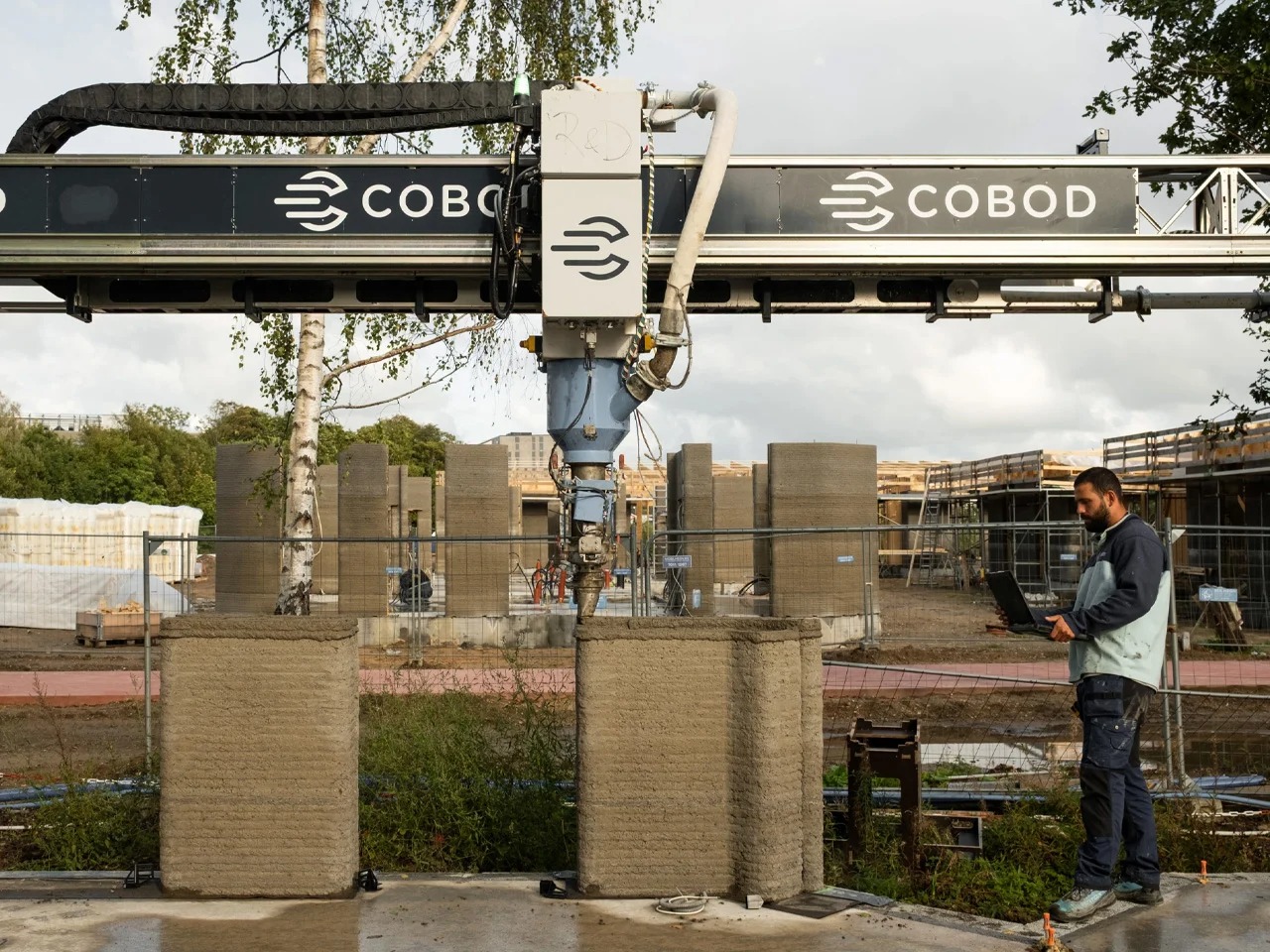 A COBOD BOD2 3D construction printer creating the concrete walls of the Skovsporet student housing project with a technician monitoring progress.