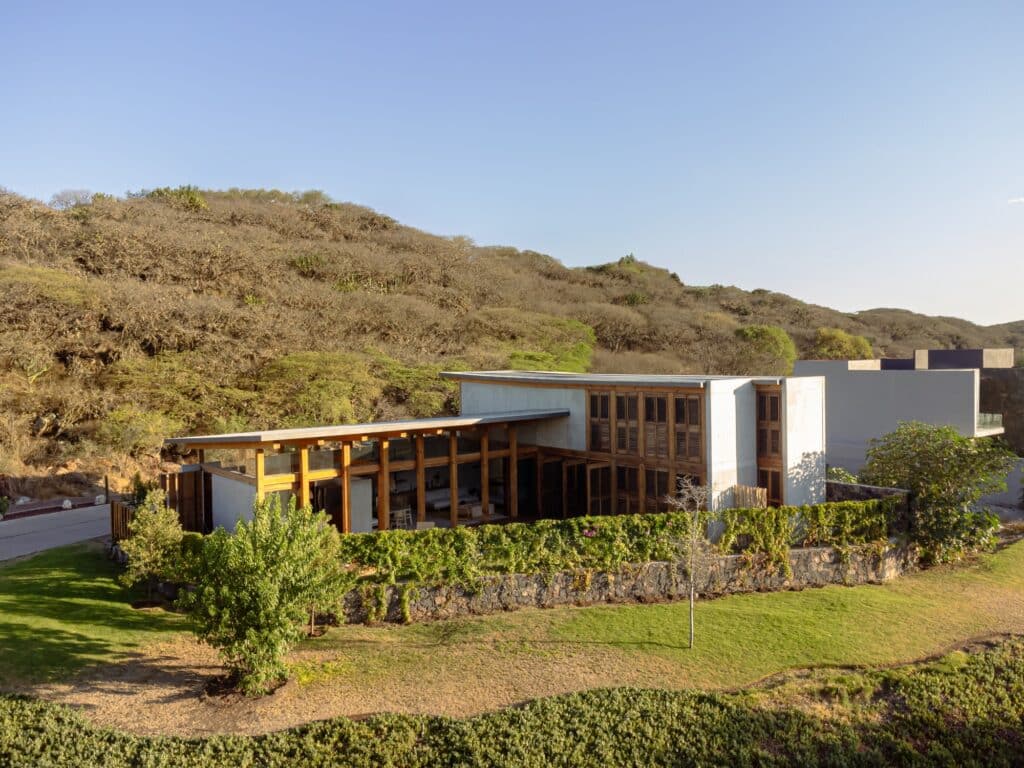 Aerial view of Queretaro House nestled against a dry forest hillside, showing its horizontal architecture and green surroundings.