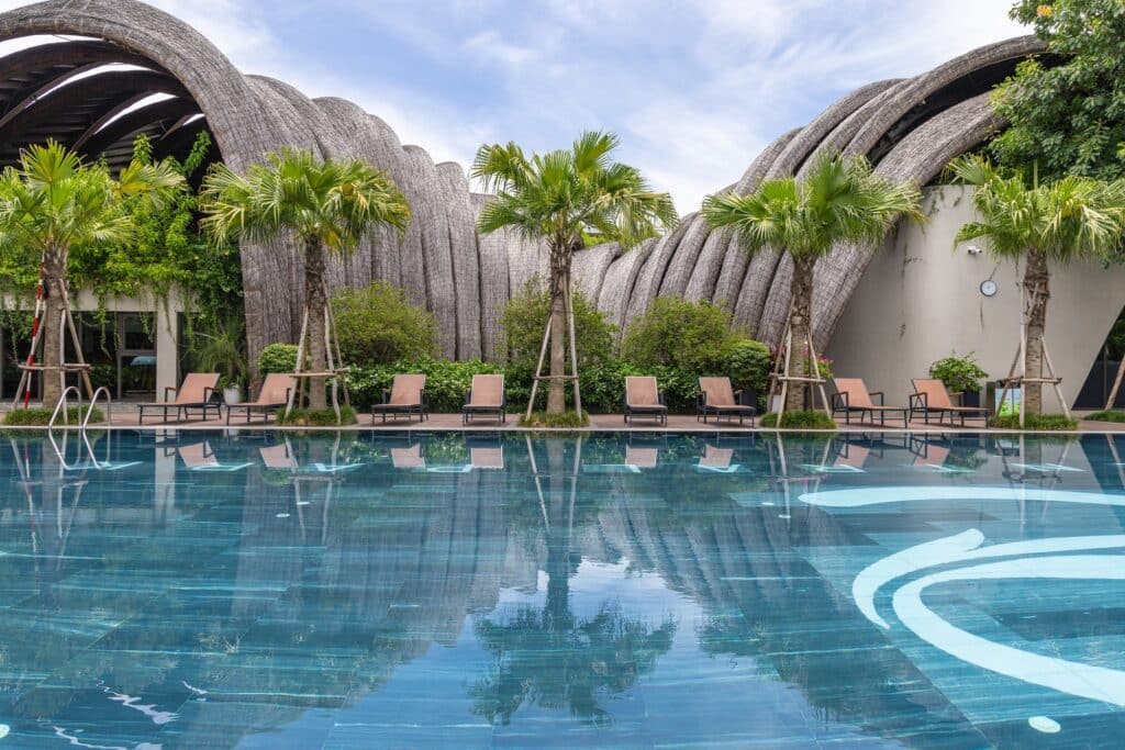 Swimming pool deck at Goot Garden House with lounge chairs and palm trees reflecting in the water against the backdrop of the arched roof.