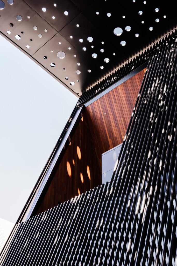 Detailed close-up of the George Street Community Building facade, showing the contrast between the black vertical metal louvers, the perforated black ceiling, and the warm interior timber cladding.
