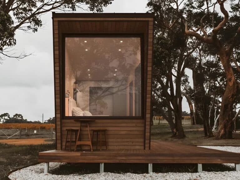 Exterior view of Ambra House, a modern tiny home with wooden cladding and a large panoramic window reflecting surrounding trees on a wooden deck.