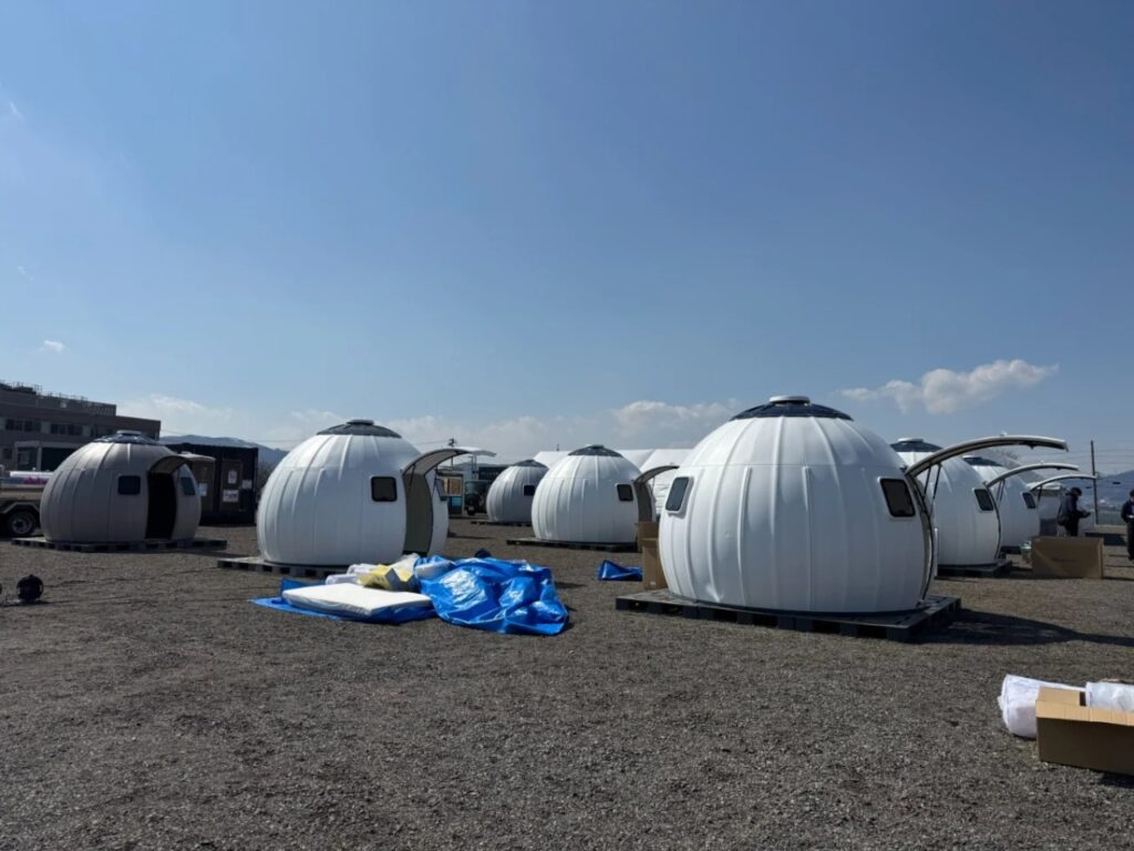 A row of white Ezdome disaster relief pods installed on a gravel site under a clear blue sky.