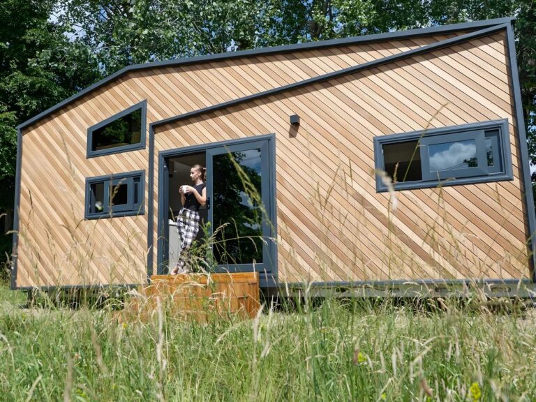 A woman standing on the porch of a modern Miami tiny home model with striking diagonal wood cladding and dark gray window frames, set against a green, grassy landscape.