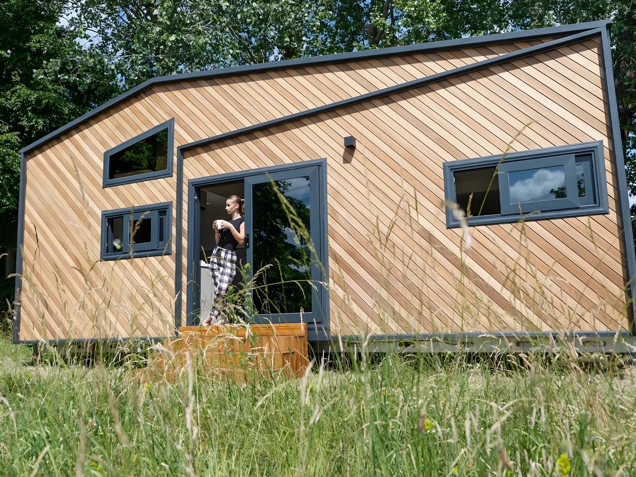 A woman standing on the porch of a modern Miami tiny home model with striking diagonal wood cladding and dark gray window frames, set against a green, grassy landscape.