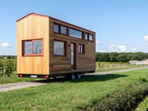 Side exterior view of 'Into the Woods' Tiny House by Baluchon on a dual-axle trailer, showcasing cedar wood cladding and red window frames in a rural setting.