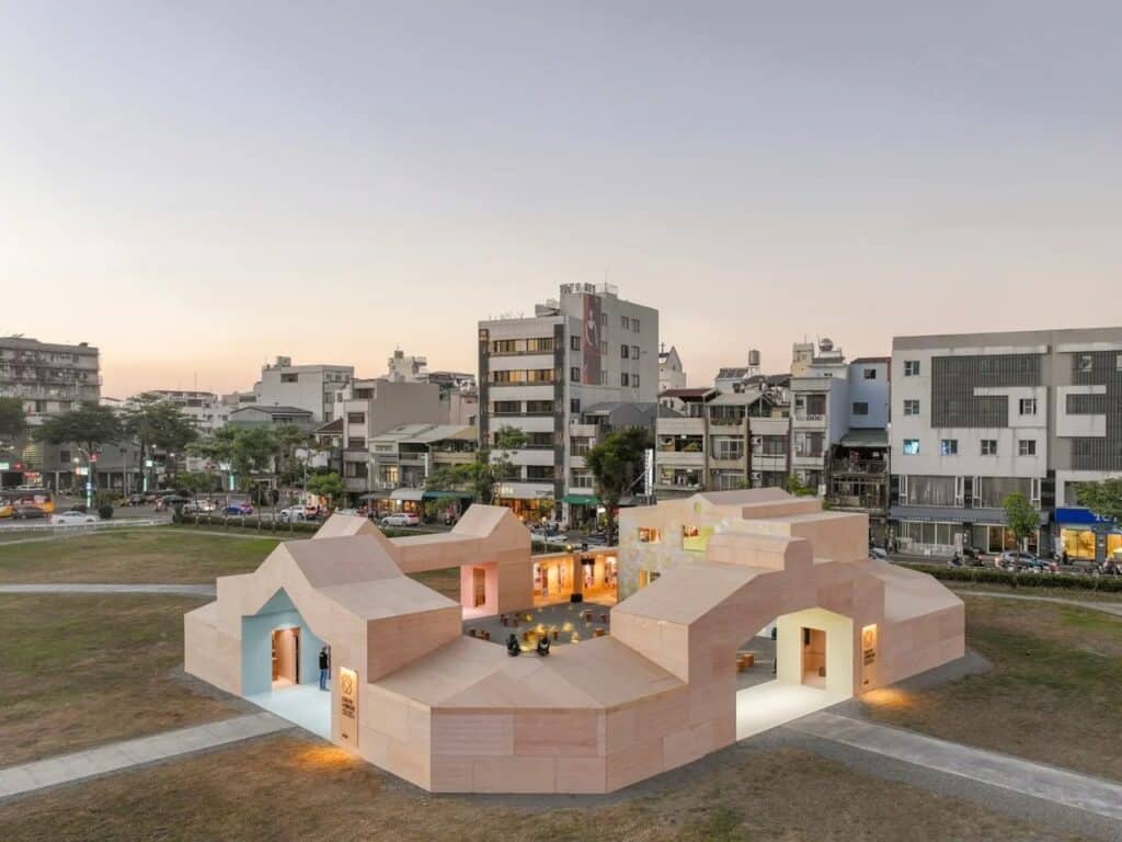Wide angle shot of the wooden pavilion at dusk with city buildings in the background and warm interior lighting reflecting on the grass.