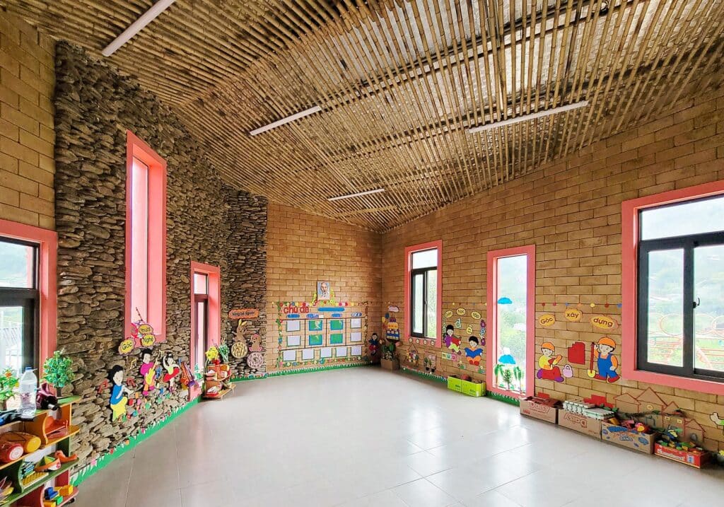 Interior classroom view with walls made of local earth bricks and stone, featuring a ceiling crafted from natural bamboo sticks and colorful educational decorations.