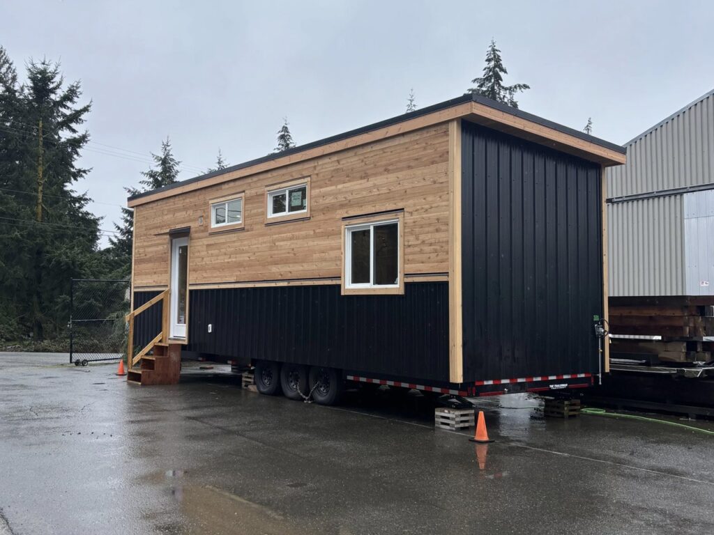 Profile view of the Juniper tiny house on a triple-axle trailer showing the contrast between timber and dark metal siding.