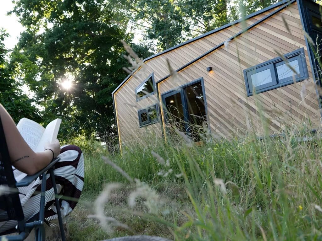 Close-up, low-angle shot of the Miami tiny house exterior with a person reading a book in a chair in the foreground, emphasizing a peaceful, sustainable lifestyle.