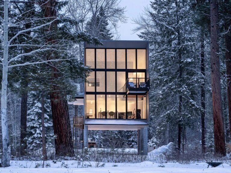 Modern, three-story Wenatchee River Cabin by Wittman Estes illuminated at dusk, set among snow-covered pine trees in the Okanogan-Wenatchee National Forest.