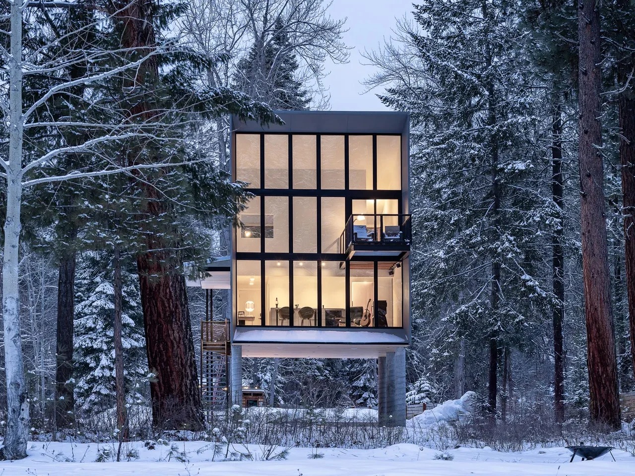 Modern, three-story Wenatchee River Cabin by Wittman Estes illuminated at dusk, set among snow-covered pine trees in the Okanogan-Wenatchee National Forest.