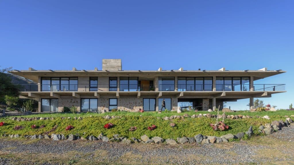 Long horizontal view of the El Drag&oacute;n house showing a rhythmic concrete structure and extensive glass facades facing the valley.