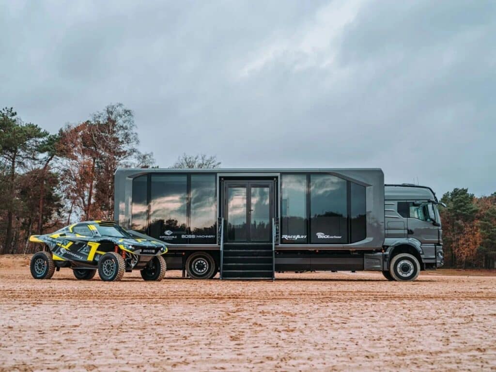 Side profile of the Shiver x Expandable unit with large glass panels, parked next to a rally car on a sandy terrain.