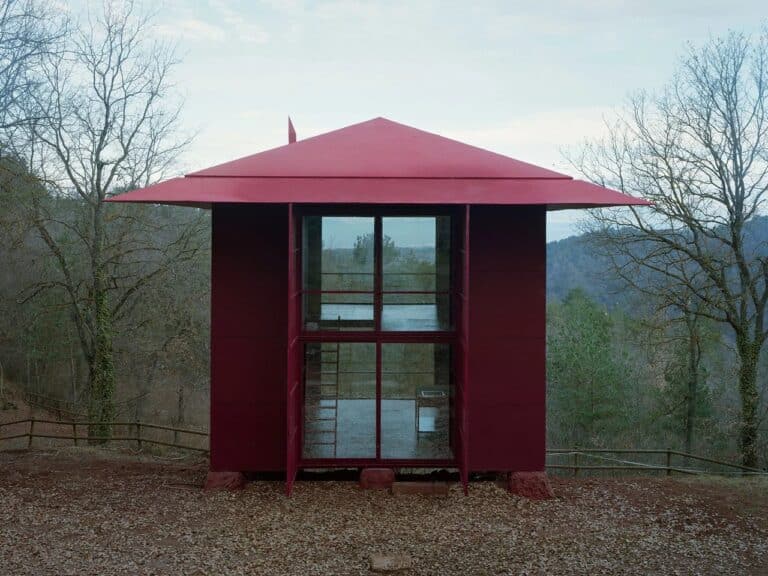 Prefab Changeover house closed facade showing modular red panels and wide eaves in a natural woodland setting.