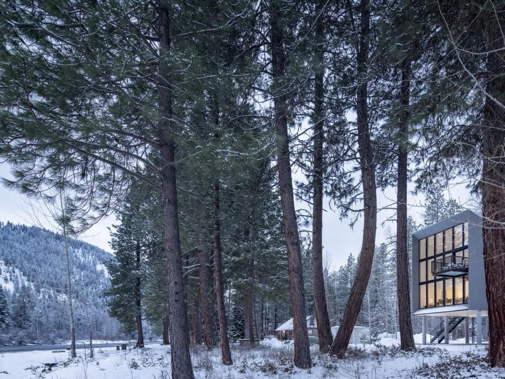 Close-up view of the minimalist, three-story Wenatchee River Cabin facade, featuring extensive grid-patterned glass windows and a small cantilevered balcony in a snowy forest setting.