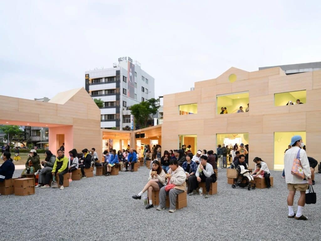 Visitors sitting on cardboard stools in the gravel courtyard of the Wonders of Wood Pavilion during a public event.