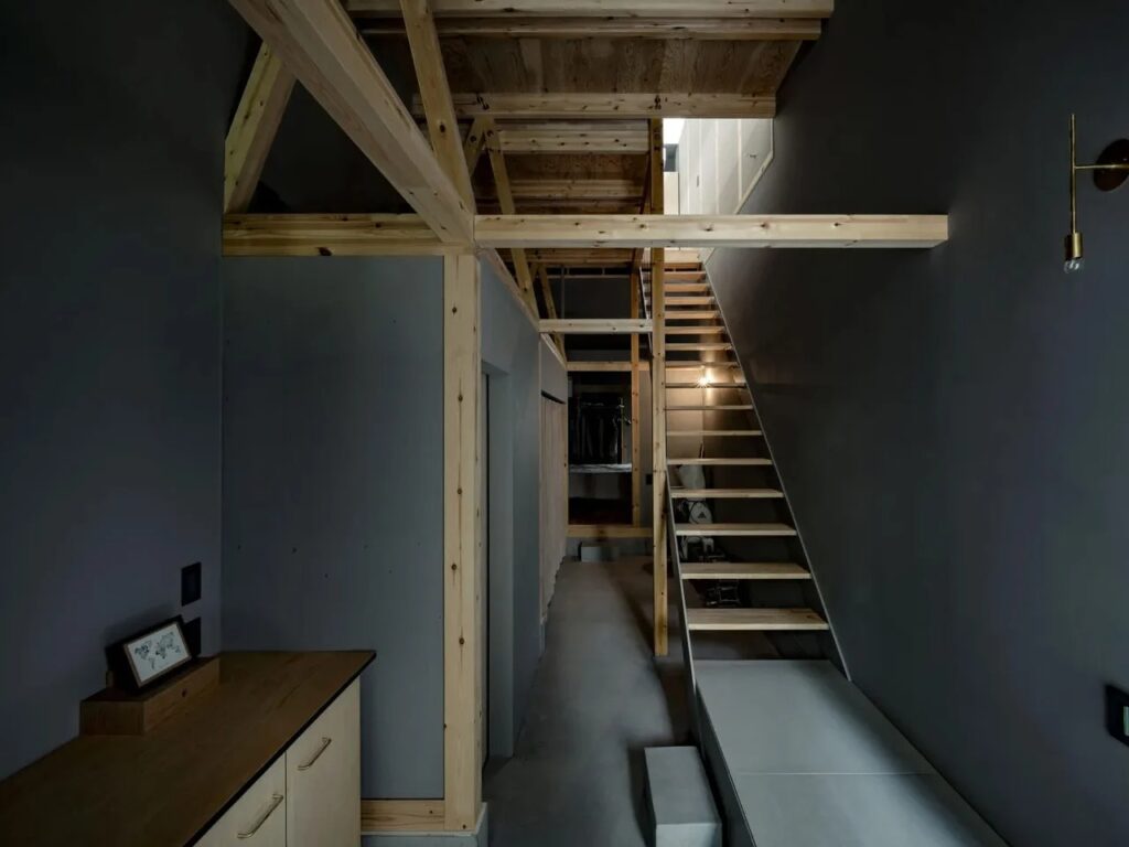 Interior view of Fukasawa House showing dark grey walls, exposed wooden timber framing, and a minimalist staircase leading to the light-filled upper floor.
