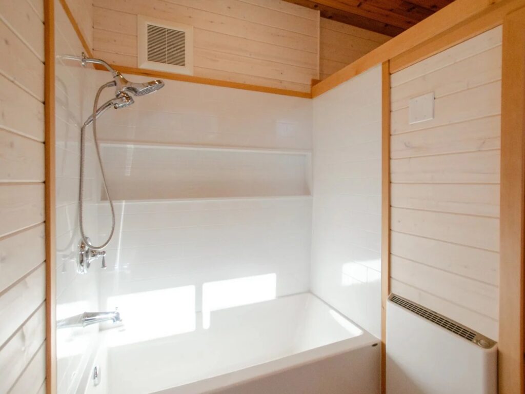 Full-sized white bathtub and shower unit inside a timber-lined tiny house bathroom.