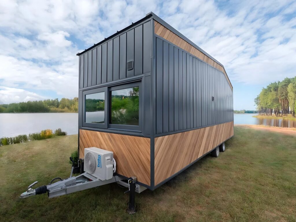 Rear three-quarter view of the mobile Miami tiny home on its trailer chassis, featuring a mix of vertical dark metal paneling and diagonal wood cladding, positioned by a tranquil lake.