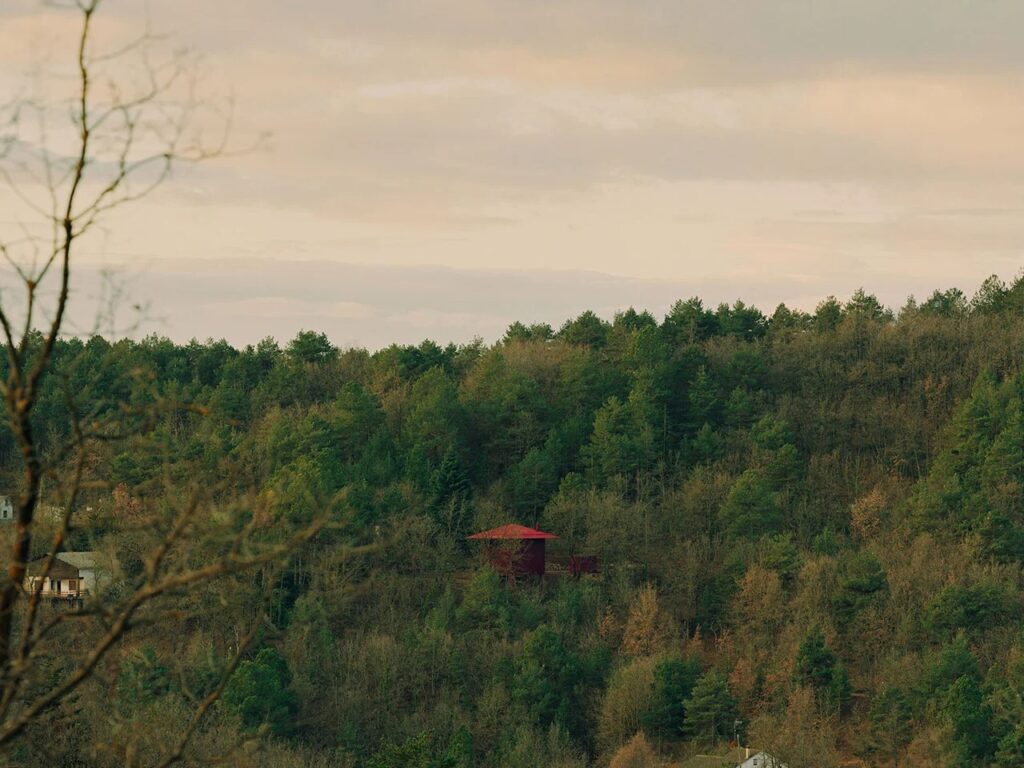 Exterior view of the Prefab Changeover House with one small square window emitting a glowing red light in a forest setting.