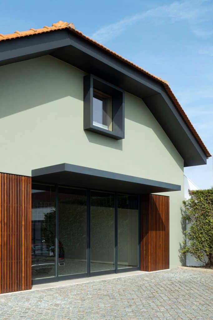 Exterior facade of Casa do Engenho featuring a sage green wall, black framed upper window, and large glass sliding doors with wooden louvers.
