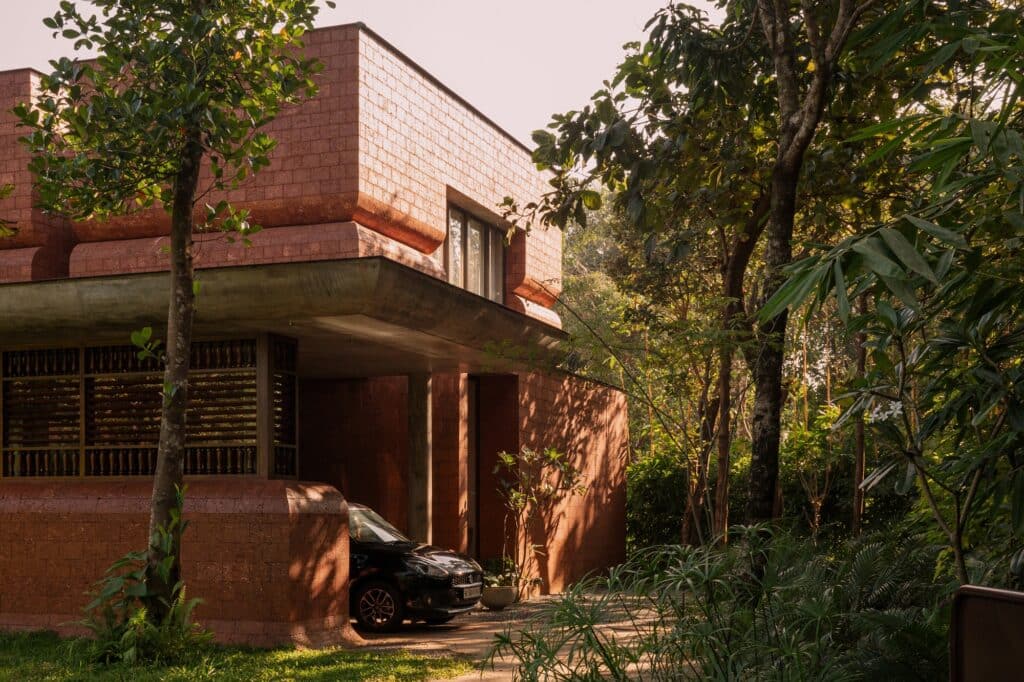 Wide elevation view of Sumesh House in Payyanur, featuring the symmetrical upper brick volumes and a deep, shaded lower level defined by timber screens, reflecting a design that integrates cultural context and outdoor space.