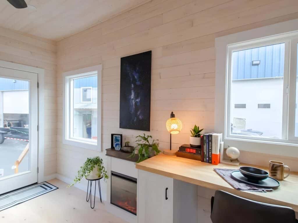 Compact workspace with a wooden desk, electric fireplace, and large windows in a tiny home.