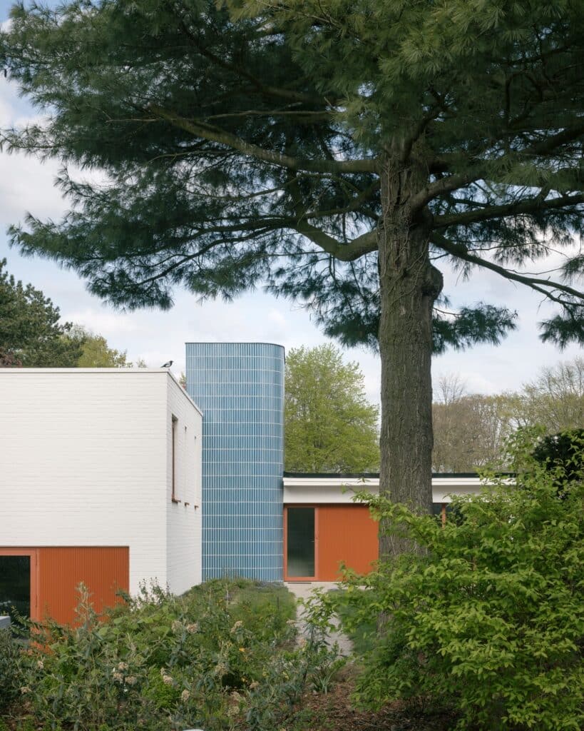 Modern bungalow seen through lush green trees, showcasing white brickwork and a distinctive blue tiled vertical volume.