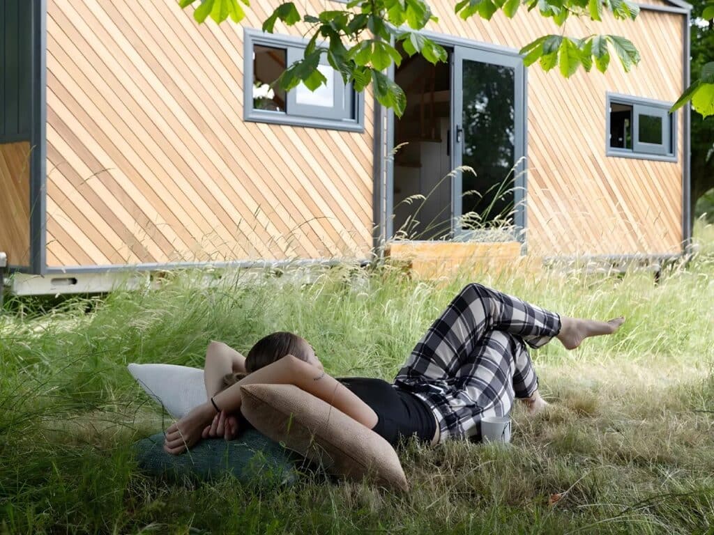 A person relaxing on the grass with pillows in front of the Miami tiny home, which has diagonal wood siding and large glass doors, promoting outdoor living.