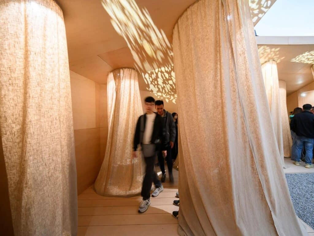 Interior view of the pavilion featuring draped linen fabrics and patterned light shadows on wooden walls with people walking through.
