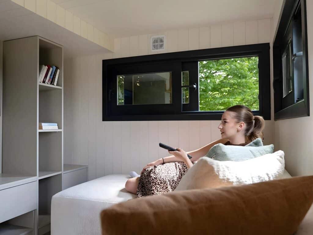 Interior shot of the Miami tiny home showing a minimalist living space with white panel walls, a bed/seating area, built-in shelving, and a large window overlooking the greenery.