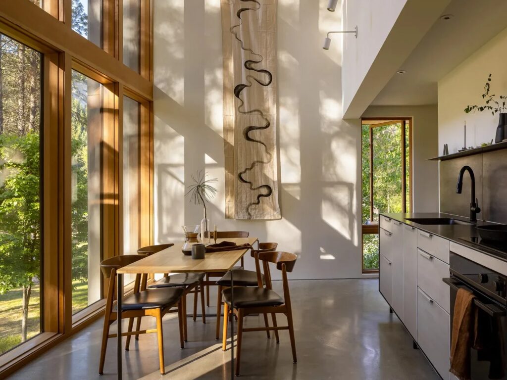 Interior view of the Wenatchee River Cabin's dining area and kitchen, featuring high ceilings, light wood window frames, a wooden dining table, and modern concrete floors.