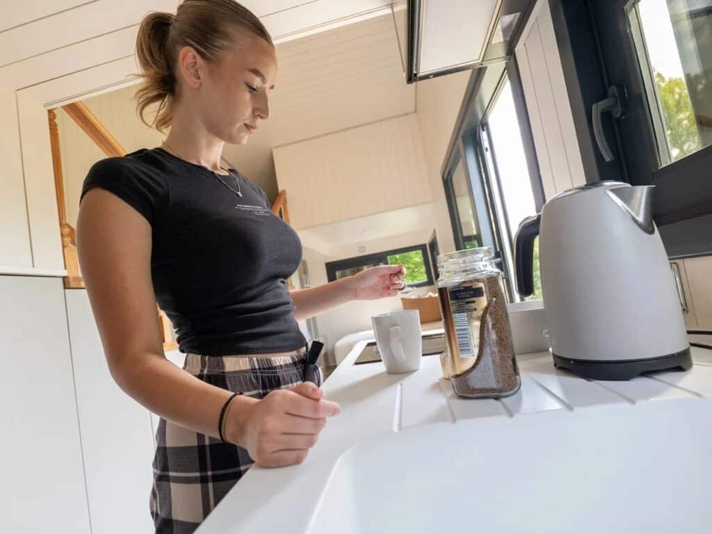 A young woman preparing a drink in the compact, contemporary kitchen of the Miami tiny home, featuring white cabinetry and a window offering natural light.