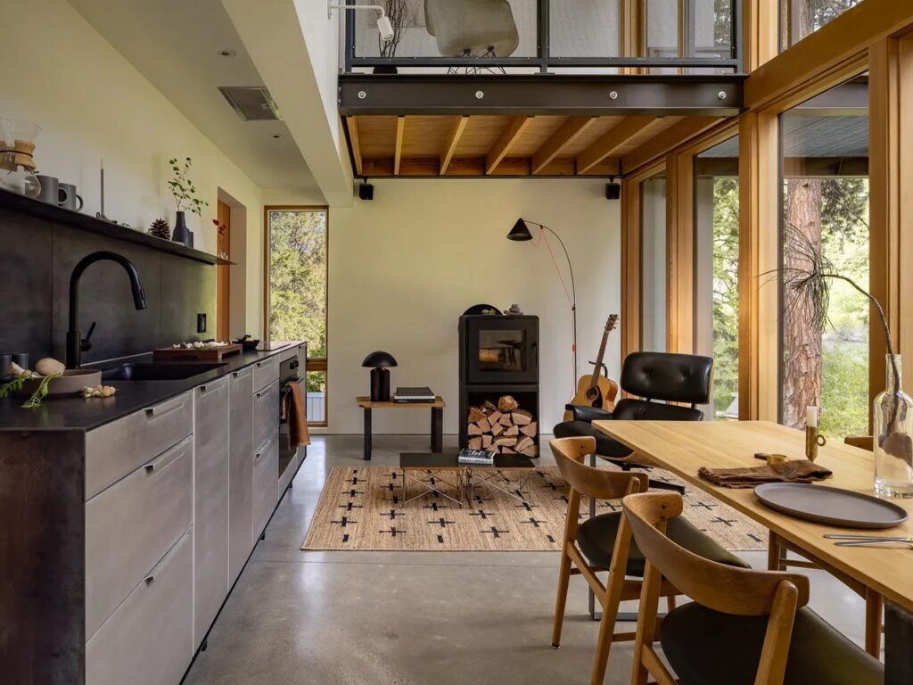 Interior view of the Wenatchee River Cabin's living area and kitchen, featuring a wood-burning stove, an Eames lounge chair, polished concrete floor, and an exposed steel beam supporting the mezzanine.