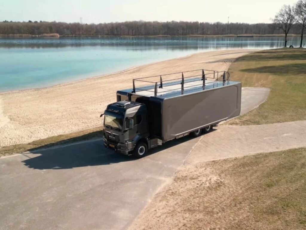 Aerial view of the Shiver x Expandable 6x6 motorhome parked by a lake, showing its sleek metallic grey exterior and rooftop terrace.