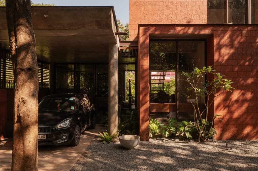 The entrance detail of Sumesh House in Payyanur showcases a functional carport covered by a concrete slab, adjacent to a wall of rich laterite brick and glazing, emphasizing the project's integration with the tropical context.