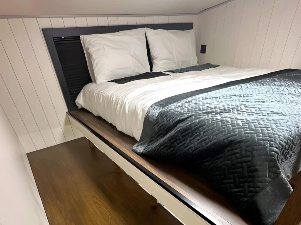 A queen-sized bed elevated on a wooden platform inside the Miami tiny home, with white and black bedding and vertical white paneling on the walls.