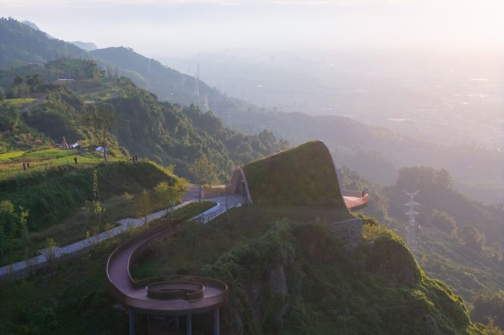 Winding mountain path leading to the Pujiang Platform with a spiral staircase in the foreground under a hazy sunset.
