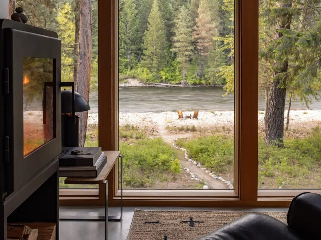 Interior view of the Wenatchee River Cabin's open-plan living and kitchen area, showing the steel-supported mezzanine, wood-burning stove, and Eames lounge chair near large windows.