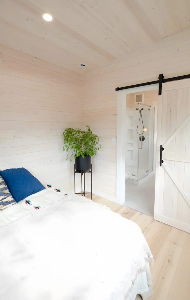 Minimalist bedroom in a tiny house with white wood walls, large egress window, and a sliding barn door for privacy.